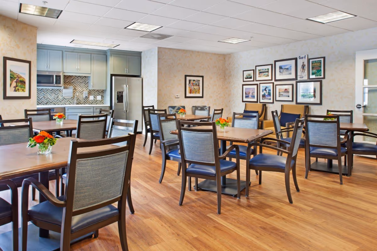 A bright dining room with multiple wooden tables and chairs arranged neatly. Each table has a small vase with orange and red flowers. The room features wood flooring, light-colored wallpaper with a subtle pattern, and a wall decorated with framed pictures. In the background, there is a kitchen area with gray cabinets, a stainless steel refrigerator, a microwave, and a patterned backsplash.