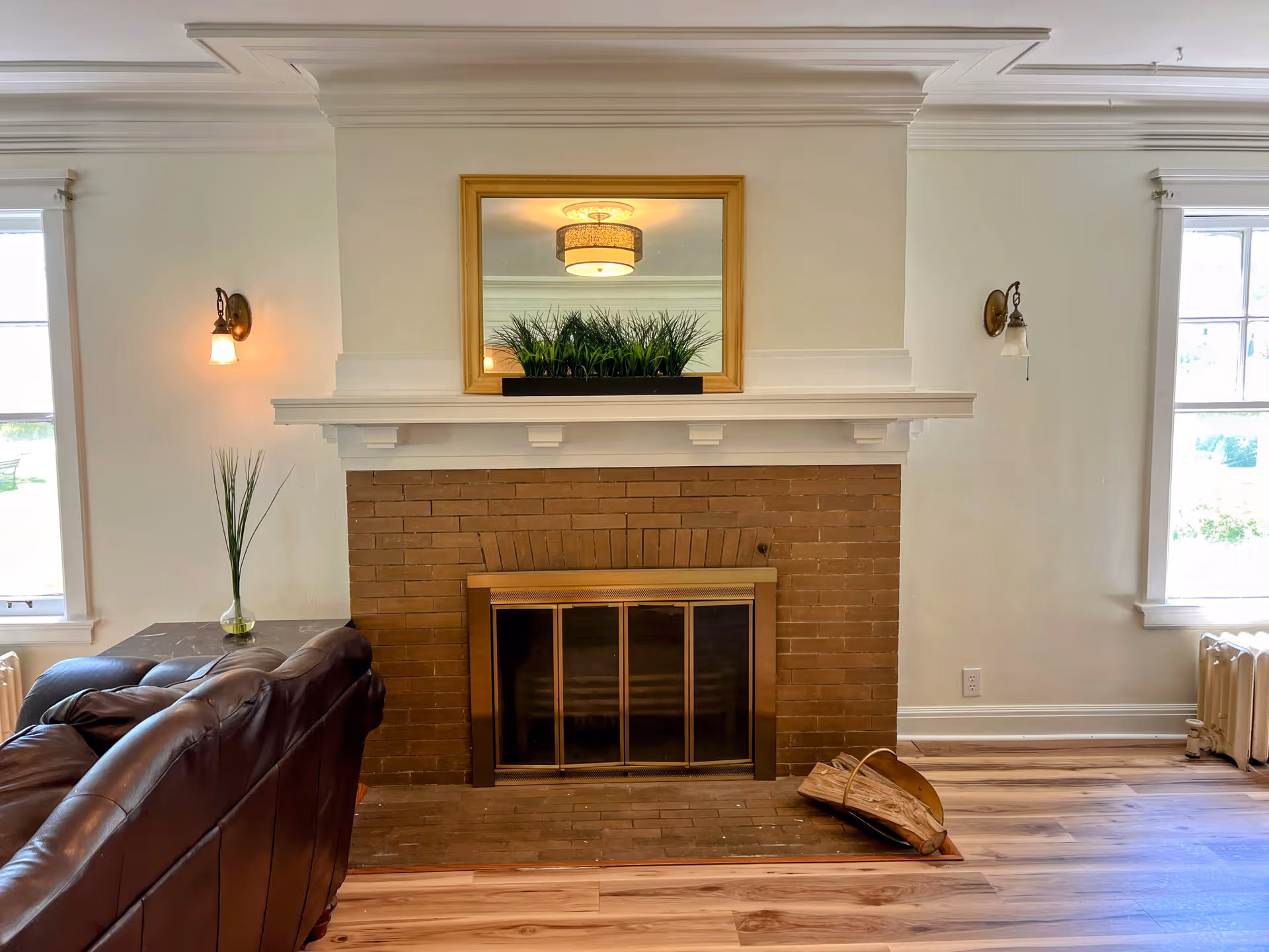 Cozy living room with a brick fireplace topped by a mirror and planter, a leather sofa, and hardwood floors.