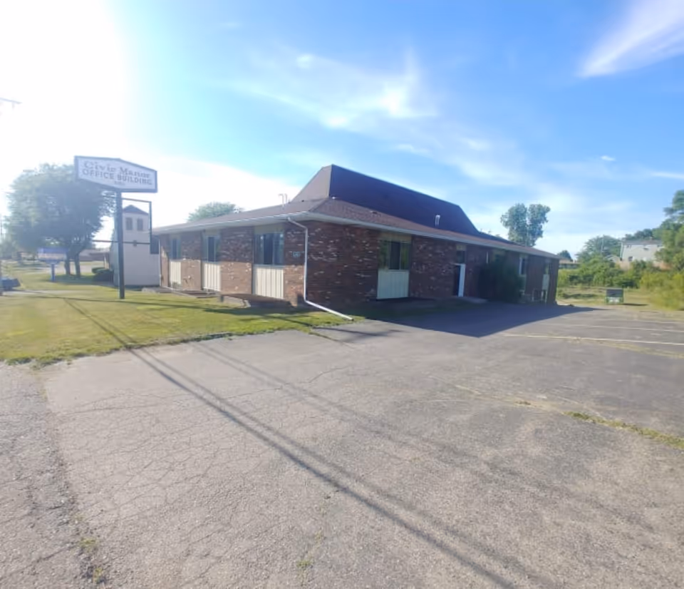 Exterior view of a single-story brick office building with a dark roof under a blue sky with some clouds. There is a sign in front of the building that reads 'Civic Manor OFFICE BUILDING'. The building is surrounded by a paved parking lot and some grassy areas with trees in the background.