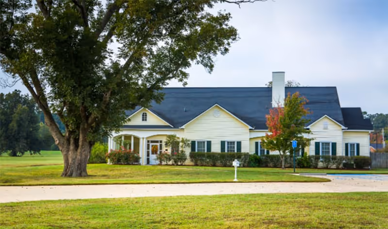 Single-story yellow residential-style building with a dark roof, lawn and a large tree in front.