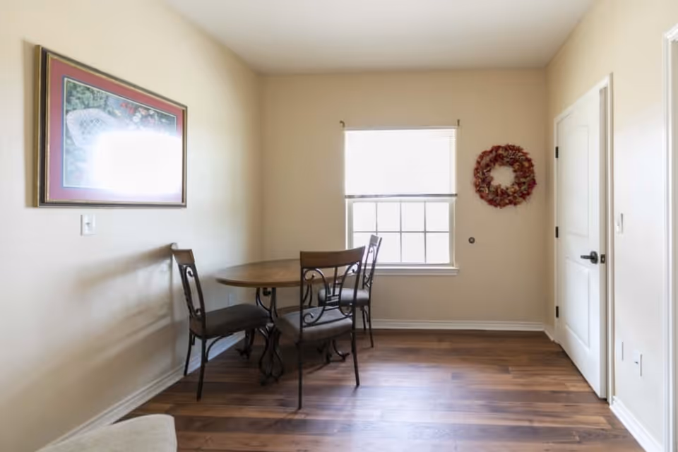 Small dining area with a round wood table, four metal-framed chairs, a window with blinds, framed wall art and a decorative wreath on beige walls.