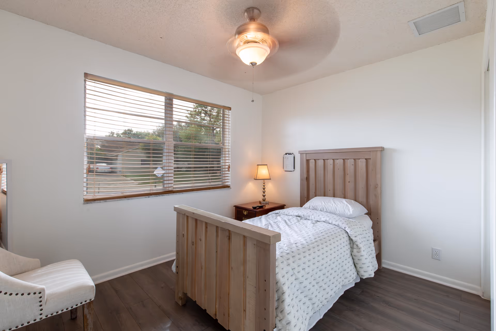 A simple bedroom with a wooden single bed covered with a white patterned blanket and pillow. Next to the bed is a wooden nightstand with a lamp and a remote control. There is a window with wooden blinds letting in natural light, a white upholstered chair with nailhead trim, and a ceiling fan with a light fixture.