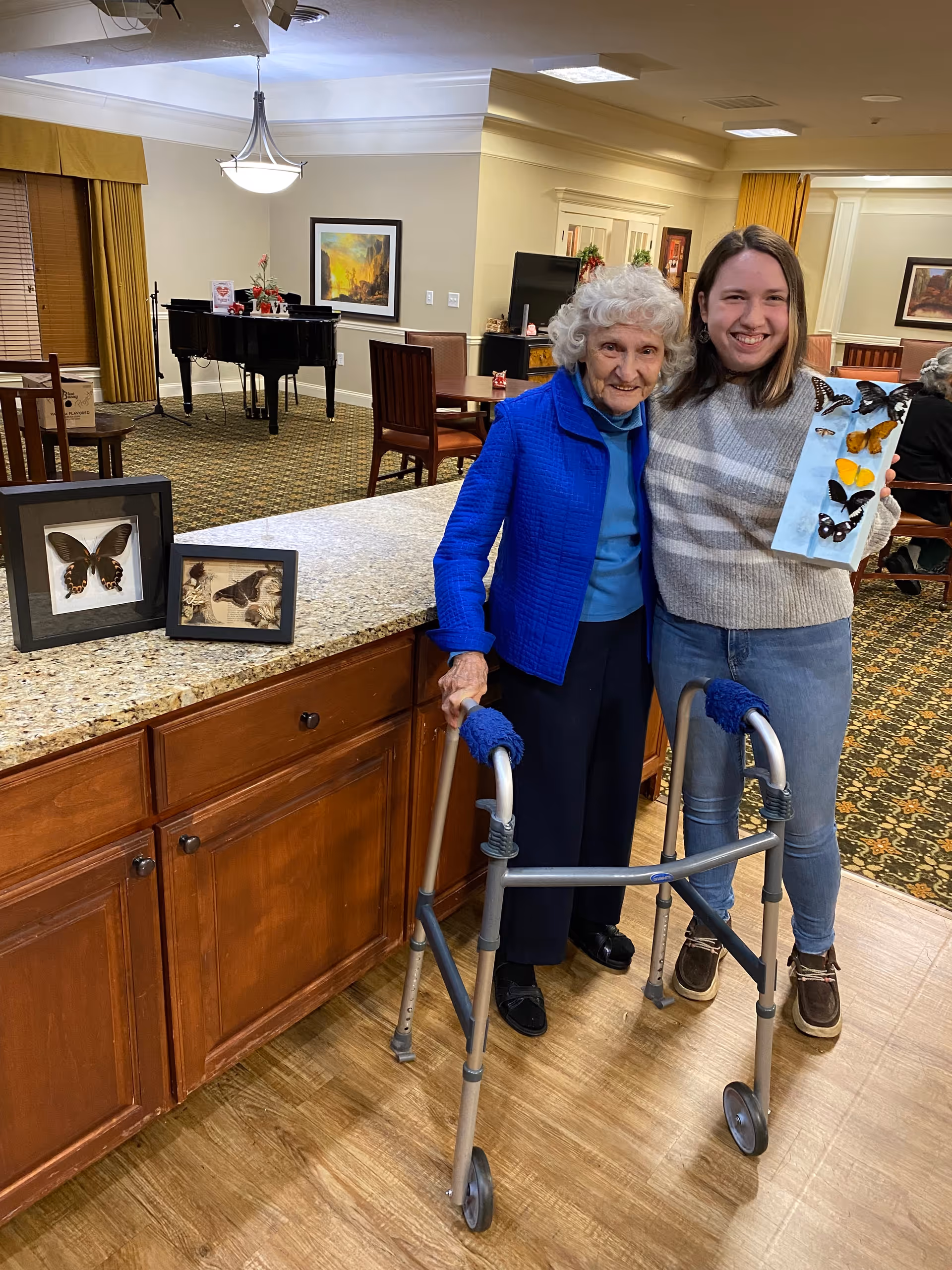 An elderly woman using a walker stands next to a younger woman holding a display of colorful butterflies. They are smiling and standing in a well-lit room with wooden cabinets, a granite countertop, and a carpeted area with chairs and a piano in the background.