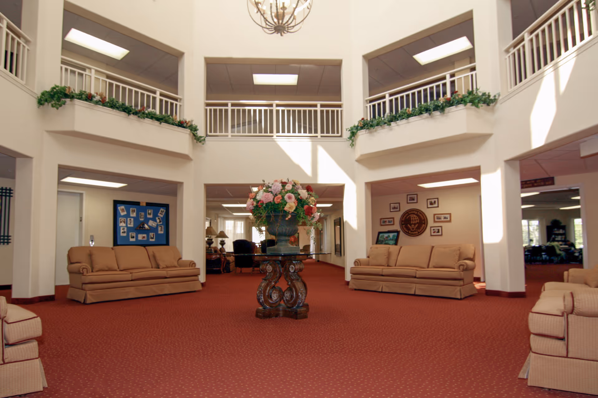 Spacious two-story common area with beige sofas arranged around the perimeter, a central decorative table with a large floral arrangement, red carpet flooring, white walls, and upper-level balconies with white railings and greenery.