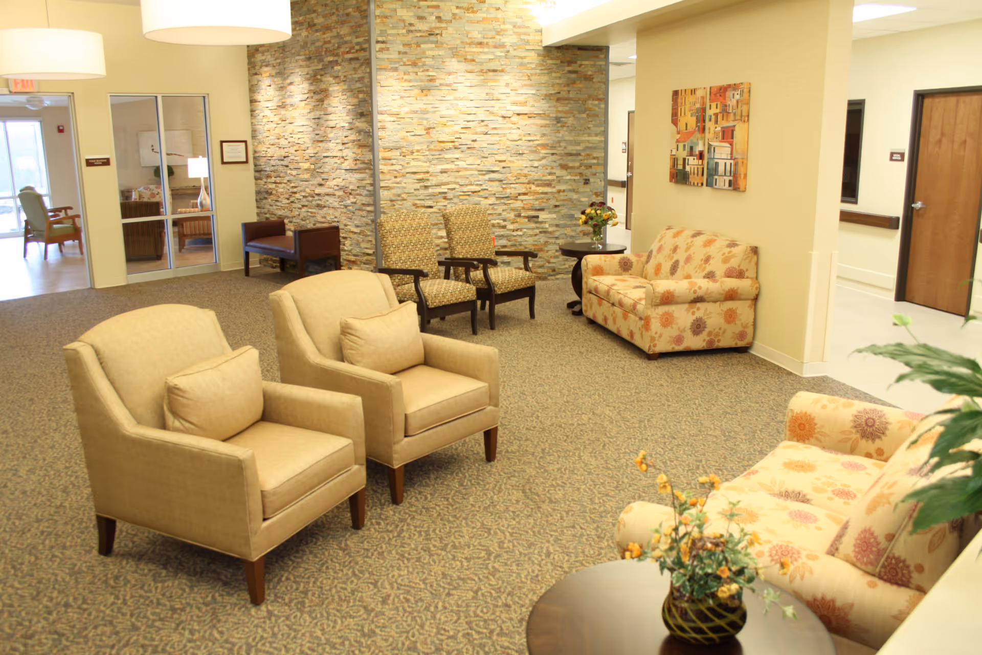 A cozy seating area in a senior living facility with beige armchairs, floral patterned sofas, a stone accent wall, a round table with a flower vase, and warm lighting fixtures.