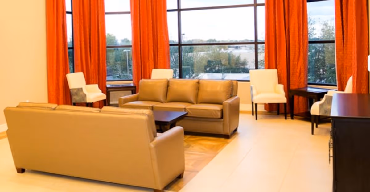 A bright living room with large windows covered by orange curtains. The room features two beige leather sofas facing each other with a dark wooden coffee table in between. There are four white armchairs placed near the windows and a dark wooden cabinet on the right side. Outside the windows, trees and buildings are visible.
