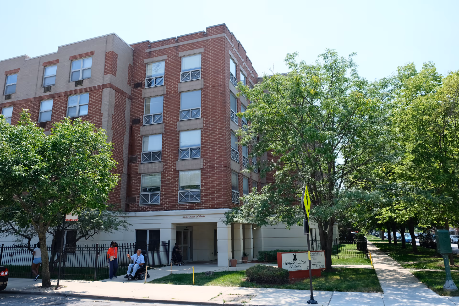 Exterior view of a multi-story brick and beige building named Senior Suites of Austin, with several windows and a main entrance. There are trees and a sidewalk in front of the building, along with a few people near the entrance, including one person in a wheelchair. A sign with the facility's name is visible on the lawn near the sidewalk.