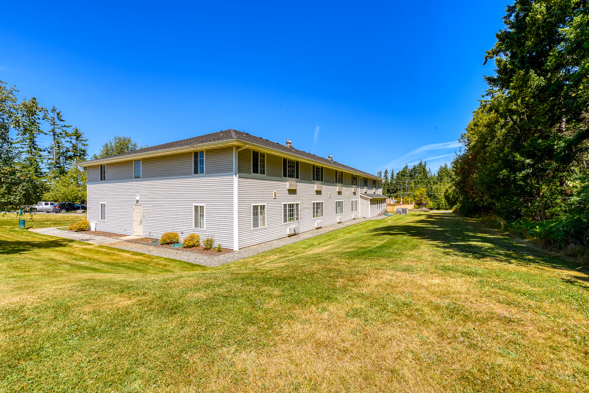 Exterior view of a two-story beige building with multiple windows, surrounded by a large grassy area and trees under a clear blue sky.