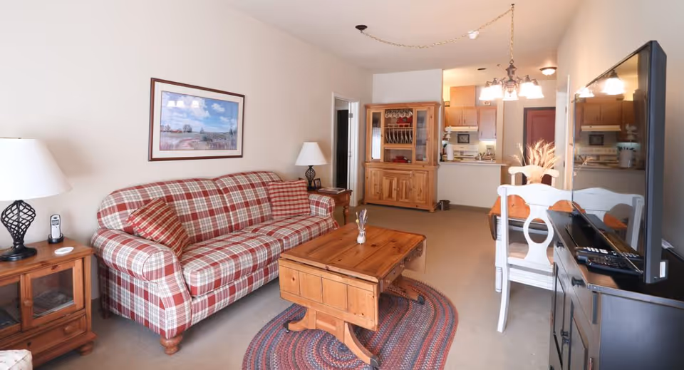 A cozy living room with a red and white plaid sofa, a wooden coffee table on a braided oval rug, a wooden side table with a lamp and phone, a wooden hutch, and a dining area with white chairs and a wooden table. The kitchen is visible in the background.