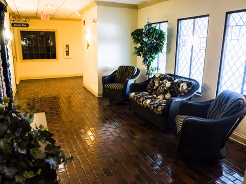 A hallway with brown tiled flooring and cream-colored walls featuring a seating area with two striped armchairs and a floral-patterned sofa. There is a potted plant next to the seating area and windows with diamond-patterned grills letting in natural light. A sign on the wall indicates the direction to the Beauty Shop.