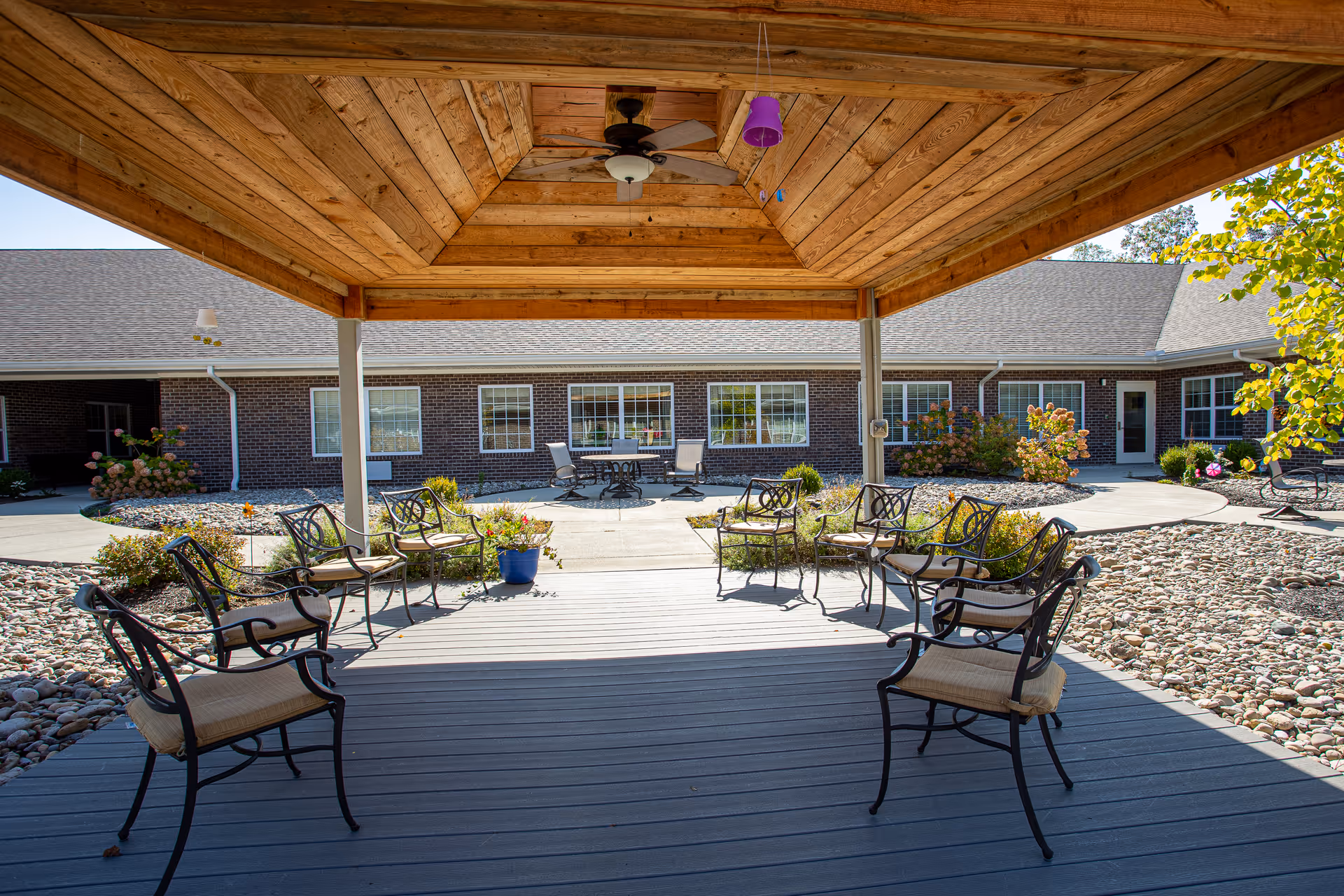 Outdoor covered seating area with metal chairs arranged in a semi-circle on a wooden deck, overlooking a courtyard with a paved walkway, rock landscaping, and a brick building with multiple windows in the background under a clear sky.