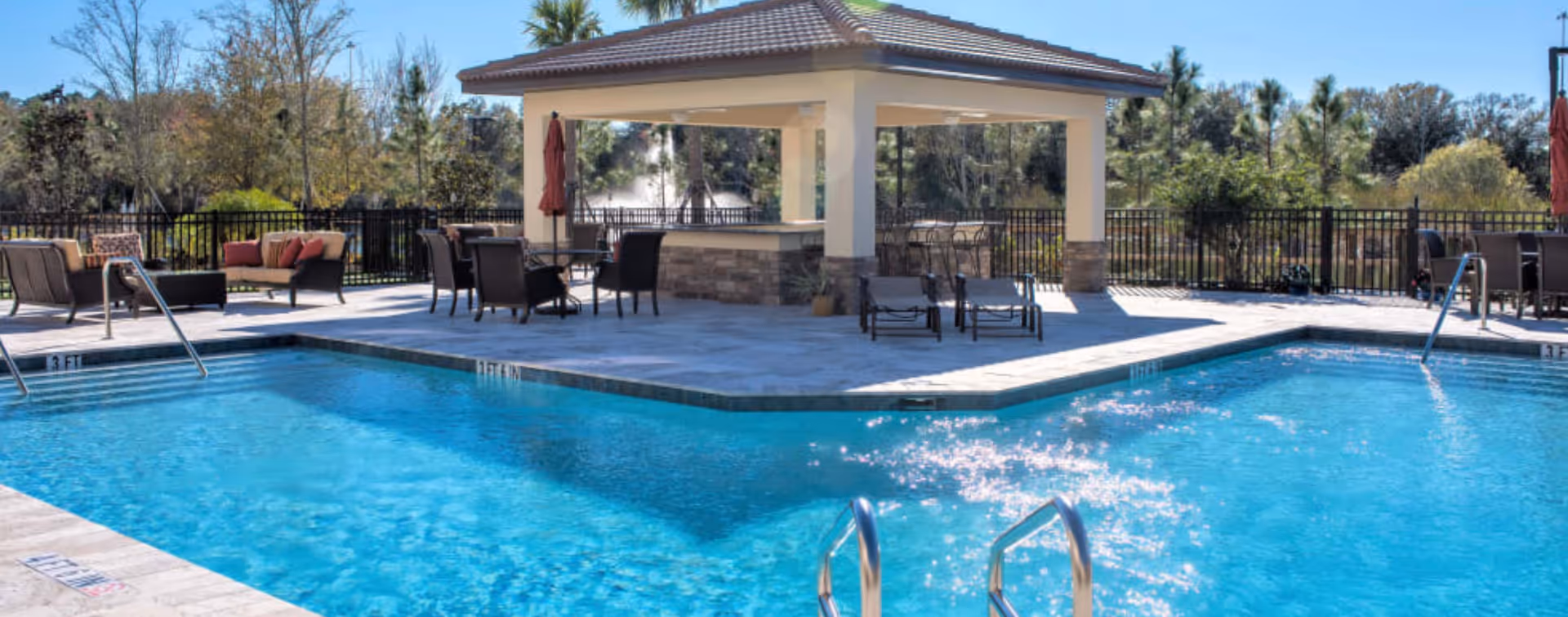 Outdoor swimming pool area with clear blue water, surrounded by a tiled deck with lounge chairs, tables, and umbrellas. A covered pavilion with seating is in the background, along with trees and a black metal fence enclosing the area.