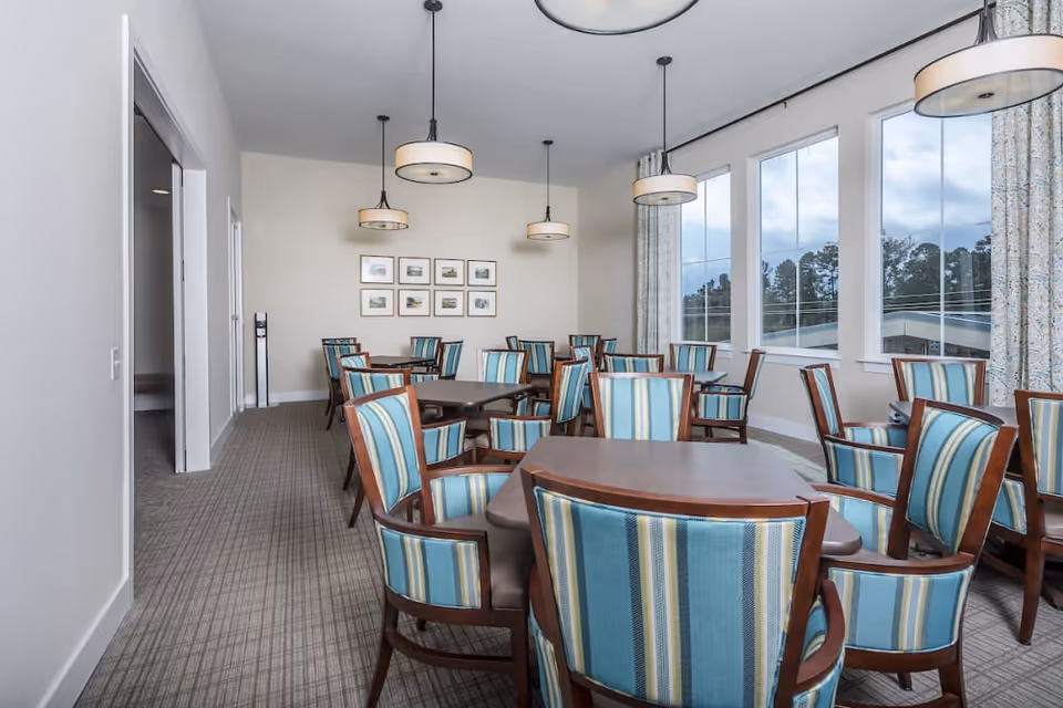 Bright communal dining room with multiple wooden tables and striped upholstered chairs beneath pendant lights and large windows.