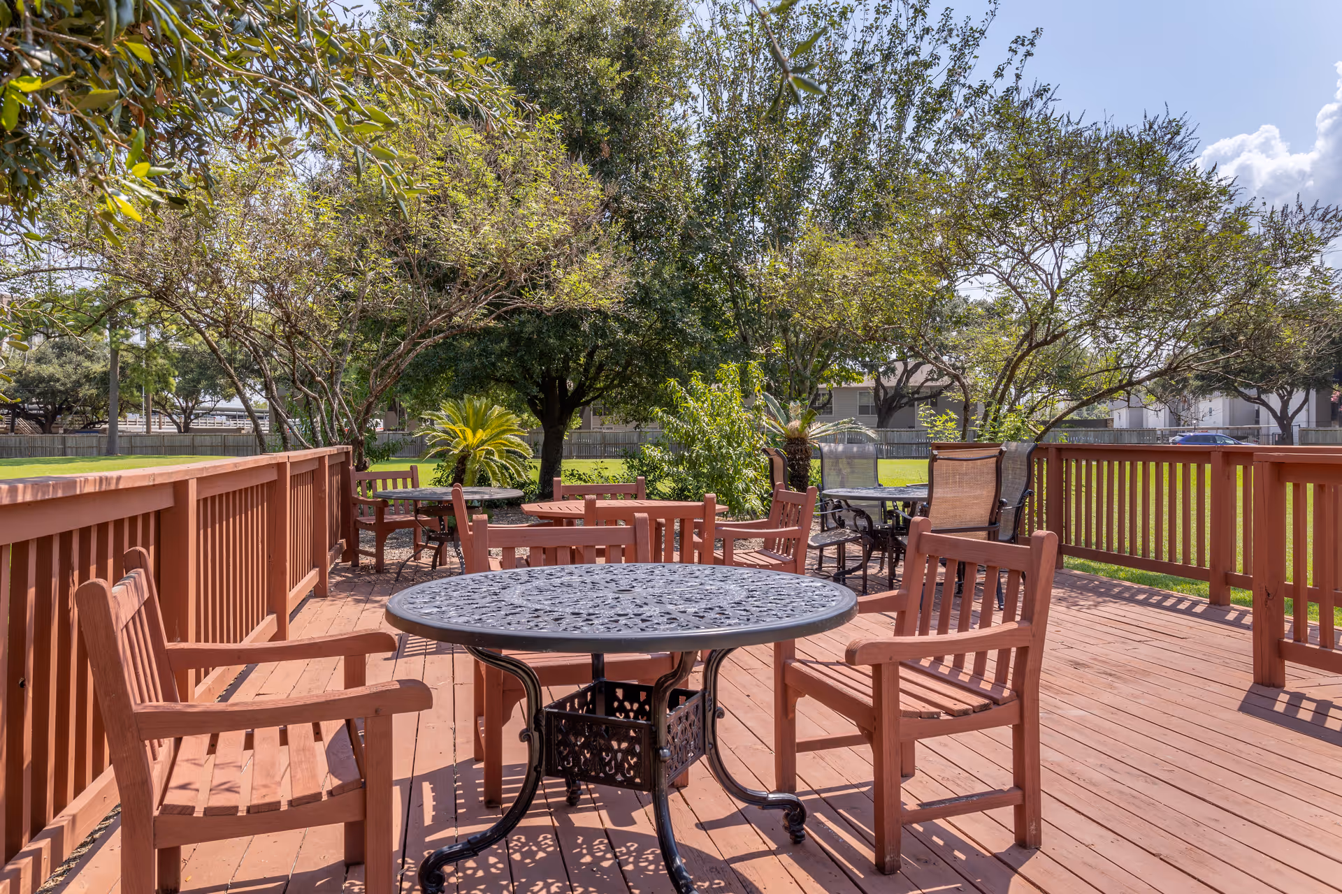 Outdoor wooden deck area with multiple tables and chairs surrounded by trees and greenery under a partly cloudy sky.