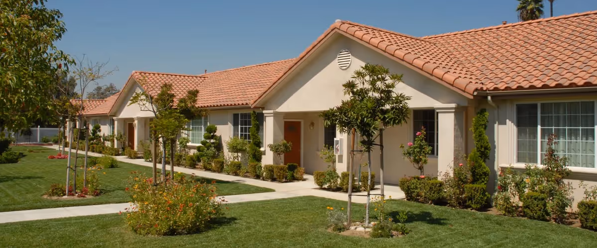 Exterior view of a single-story retirement center building with beige walls and a red tiled roof. The building is surrounded by well-maintained green lawns, small trees, and shrubs under a clear blue sky.