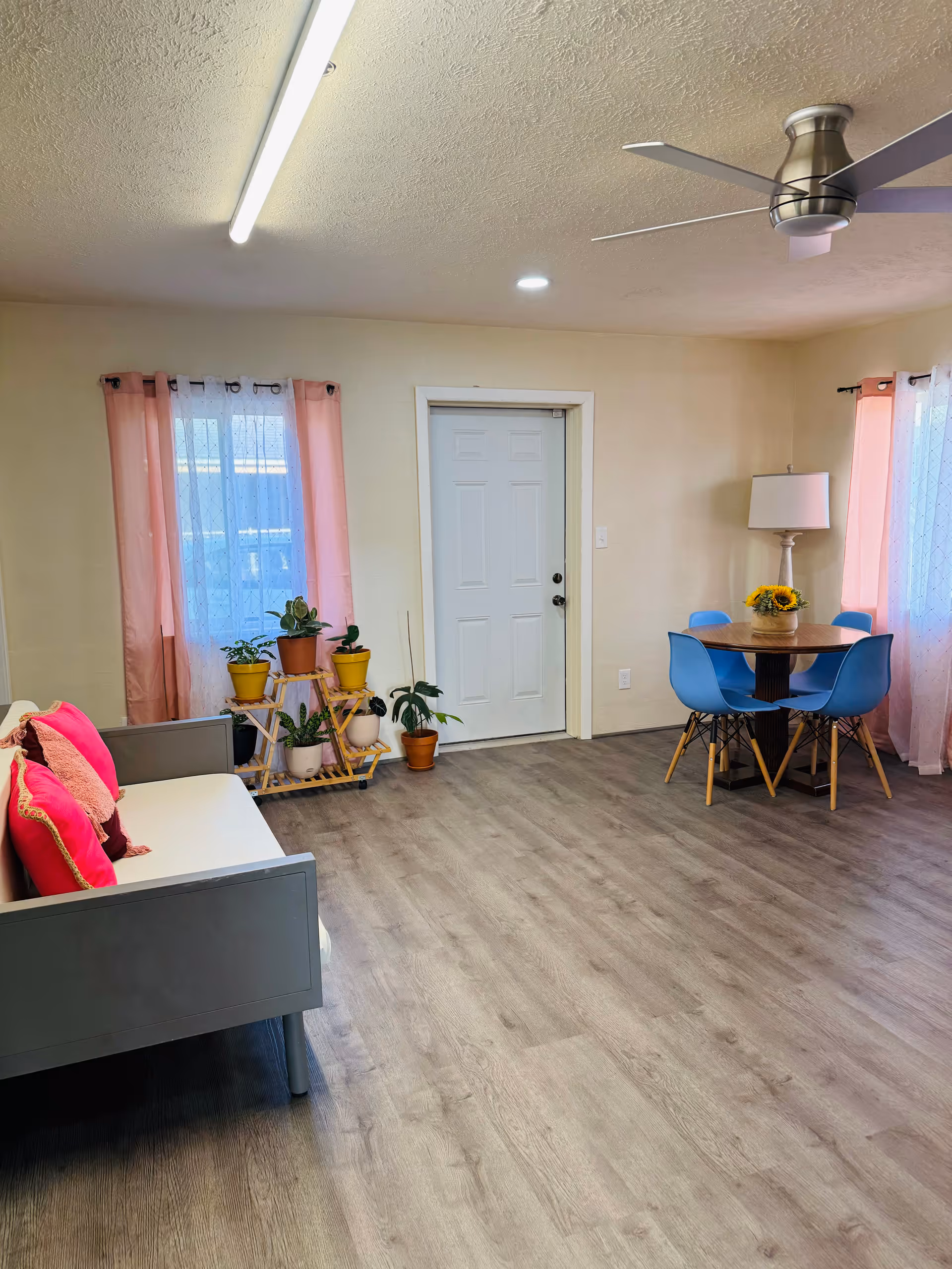 A bright and clean living room area with light wood flooring, a gray sofa with pink cushions on the left, a small wooden plant stand with several potted plants near a window with pink curtains, a white door in the center, and a round wooden dining table with four blue chairs and a sunflower centerpiece on the right. A floor lamp stands next to the dining table, and a ceiling fan with lights is visible on the ceiling.