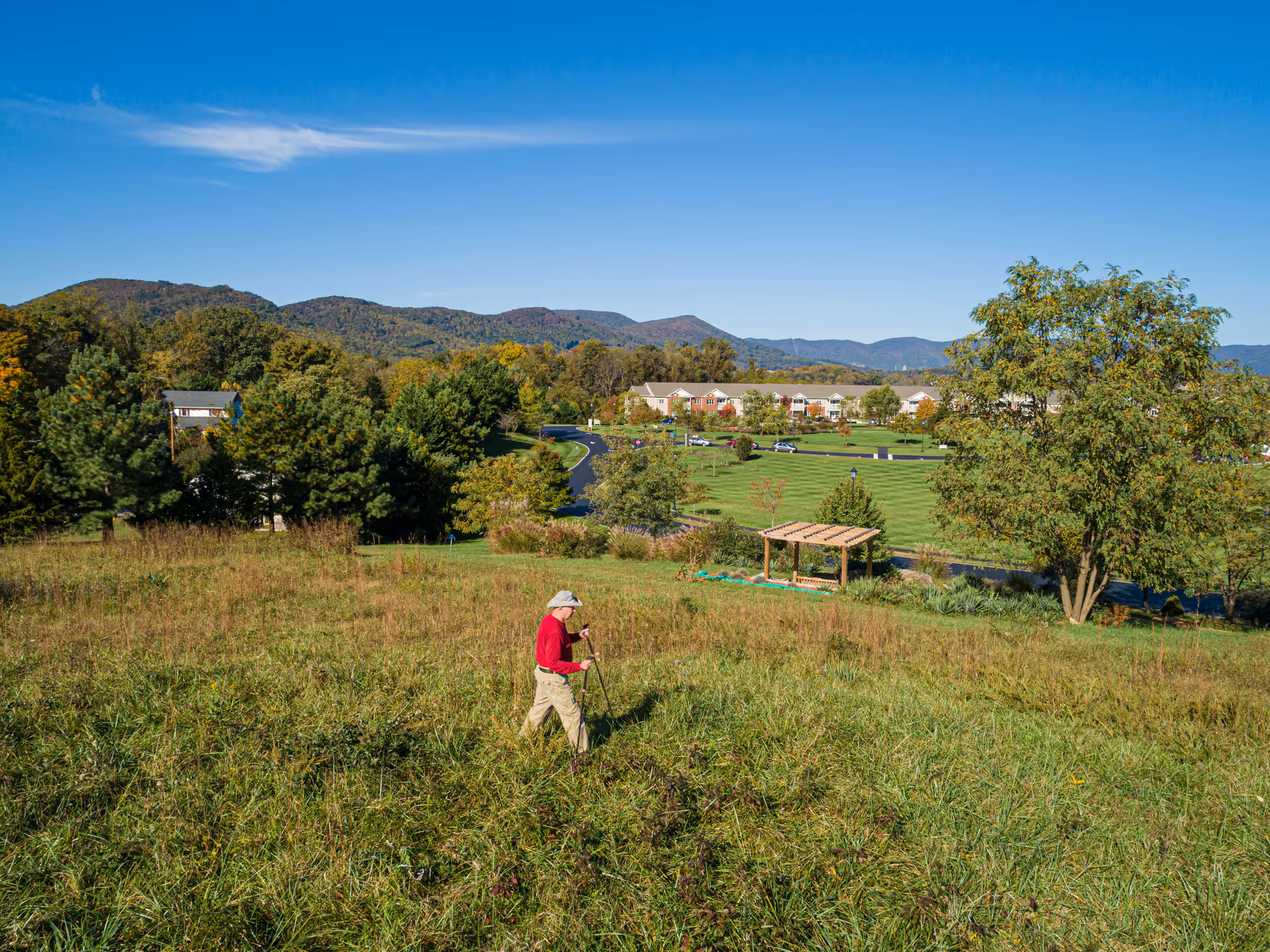 A person wearing a red shirt and beige pants walks with hiking poles through a grassy field near a retirement community. In the background, there are trees, a small wooden pergola, a road, parked cars, and buildings with mountains under a clear blue sky.