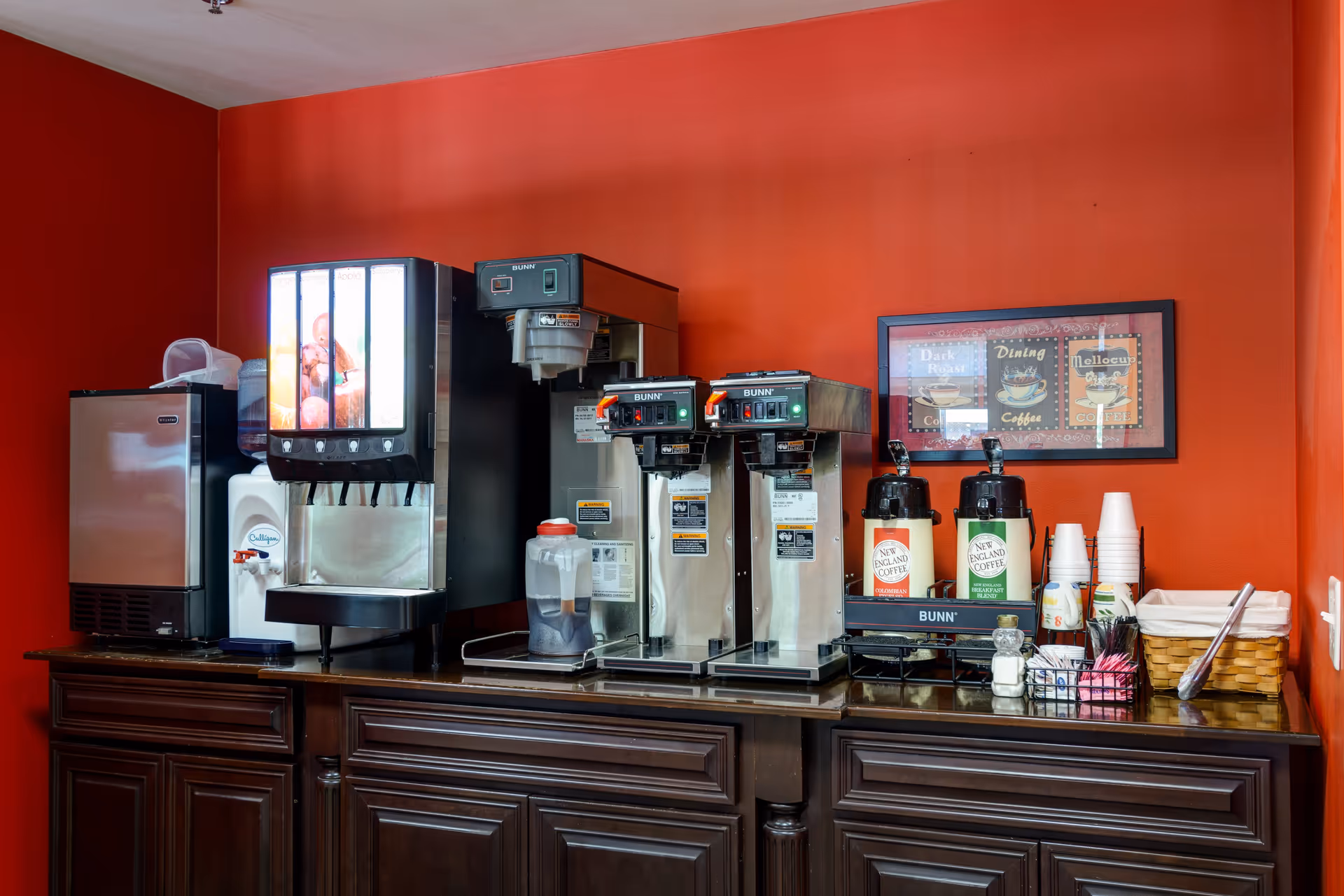 Beverage and coffee station with multiple dispensers, coffee brewers, cups and condiments on a dark wooden counter against a red wall.