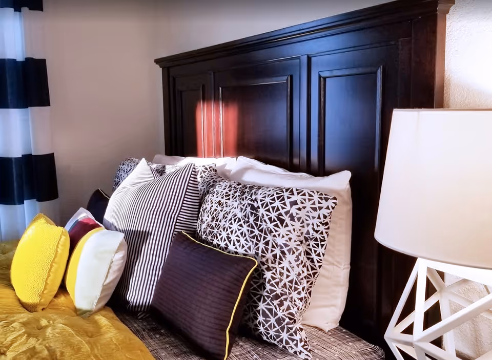 Close-up of a styled bed with multiple patterned pillows, a dark wooden headboard, striped curtains, and a bedside lamp.