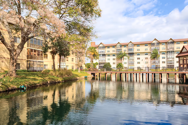 View of a senior living facility with multi-story buildings surrounding a calm pond. There is a wooden bridge crossing the pond, trees with some autumn foliage, and a partly cloudy sky above.