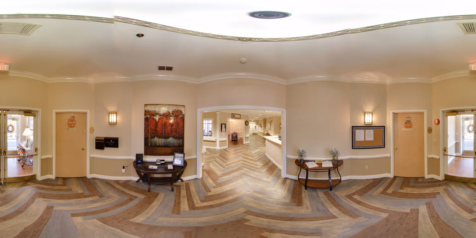 A spacious, well-lit interior hallway of a senior living facility with light beige walls and a patterned wooden floor. There are two small tables against the walls, one with a painting of red trees above it and the other with some decorative items and papers. Two doors with owl-themed decorations are visible on either side, and double glass doors lead to another area. The hallway opens into a larger corridor with more seating and a reception or nurse's station in the background.