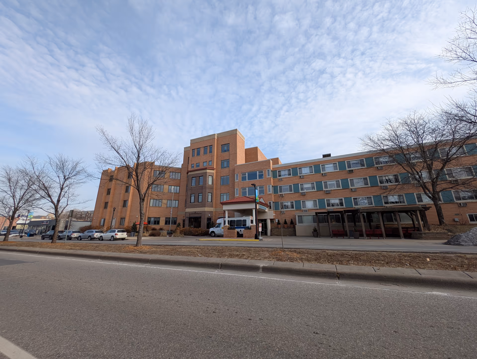 Front exterior of a multi-story brick senior care facility with parked cars and leafless trees along the street under a partly cloudy sky.