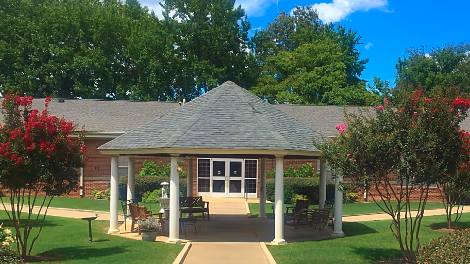 Outdoor gazebo with a gray shingled roof supported by white columns, surrounded by green grass, flowering trees, and shrubs, with a brick building and double glass doors in the background under a blue sky with some clouds.