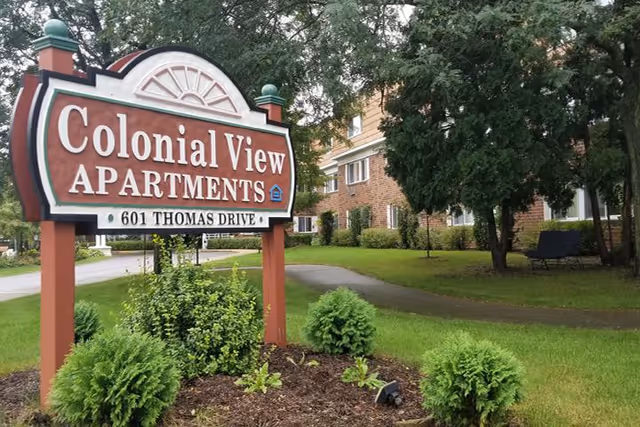 A wooden sign for Colonial View Apartments at 601 Thomas Drive stands in a landscaped area with green shrubs and grass. Behind the sign, there is a sidewalk, trees, and a brick apartment building partially visible.