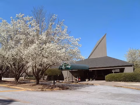 Exterior view of Woodland Ridge Assisted Living facility with a peaked roof, surrounded by blooming trees and bushes under a clear blue sky.