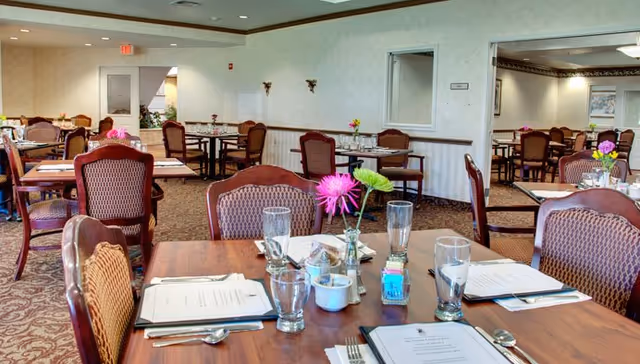 A dining room with multiple wooden tables and chairs arranged neatly. Each table has menus, drinking glasses, and small flower vases with colorful flowers. The room has carpeted floors, light-colored walls, and ceiling lights. There is a doorway and a window visible on the far wall.