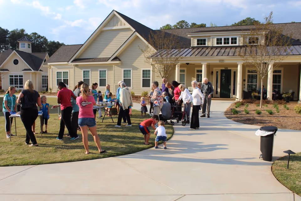 A group of people, including elderly individuals and children, gathered outside a senior living community building on a sunny day. Some people are standing near tables set up on the grass, while others are walking on the paved pathway leading to the entrance of the building. The building has beige siding, multiple windows, and a covered porch with columns.