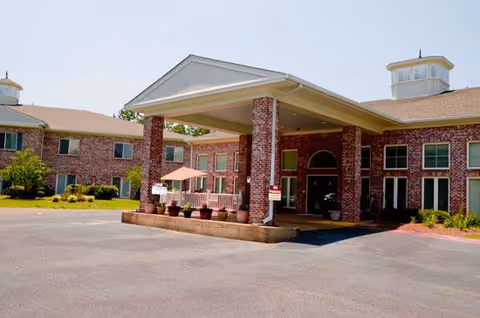 Front exterior view of a senior living facility named Castlewoods Place featuring a large covered entrance supported by brick columns, red brick building facade, multiple windows, and a clear blue sky.