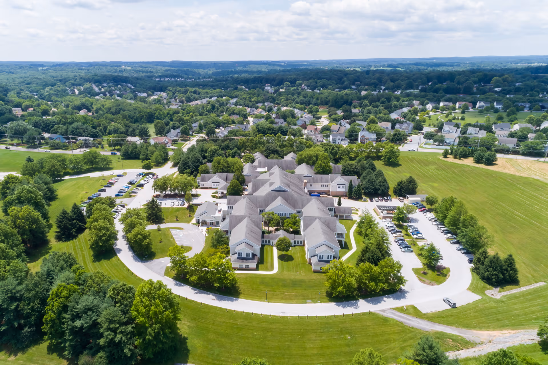 Aerial view of a large senior living facility named Copper Ridge surrounded by green lawns, trees, and parking lots, with a residential neighborhood and forested hills in the background under a partly cloudy sky.