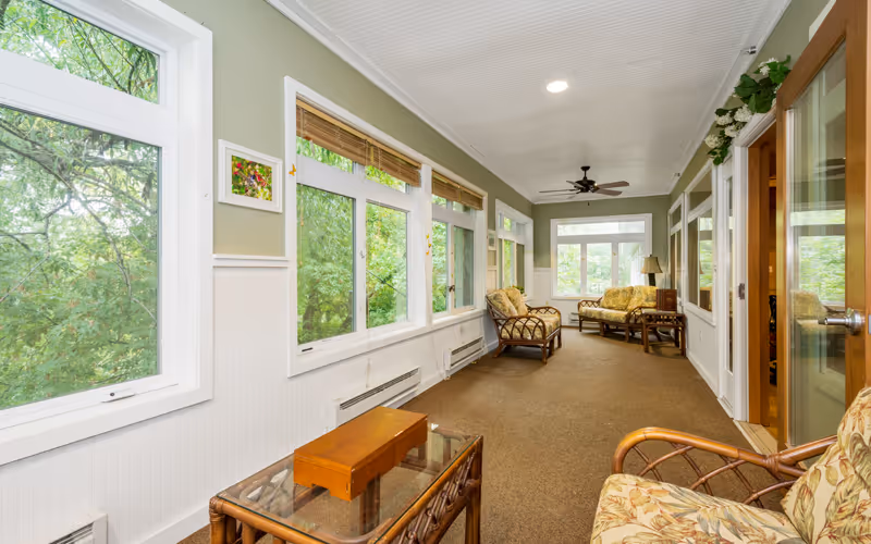 A bright sunroom with large windows on one side showing green trees outside. The room has wicker furniture with floral cushions, including chairs and a loveseat, a glass-top coffee table, and a ceiling fan. The walls are painted light green with white wainscoting, and there is a door with glass panels on the right side.