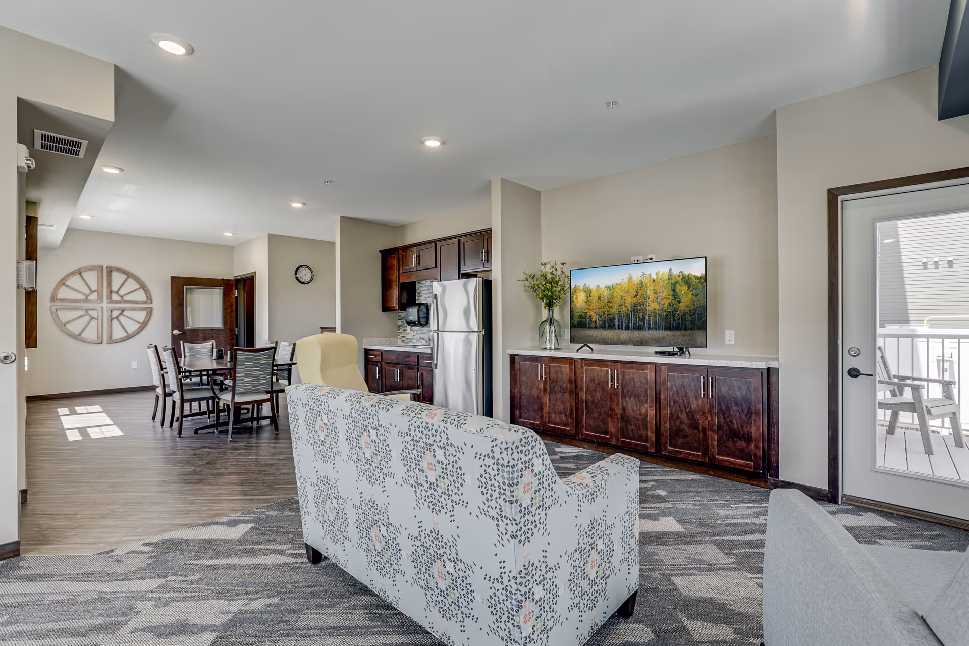 A bright and spacious living area in a senior living facility featuring a patterned sofa and armchair facing a flat-screen TV mounted on a wall above a wooden cabinet. To the left, there is a dining area with a table and chairs, and a small kitchen area with dark wood cabinets and stainless steel appliances. A glass door leads to an outdoor patio with chairs.