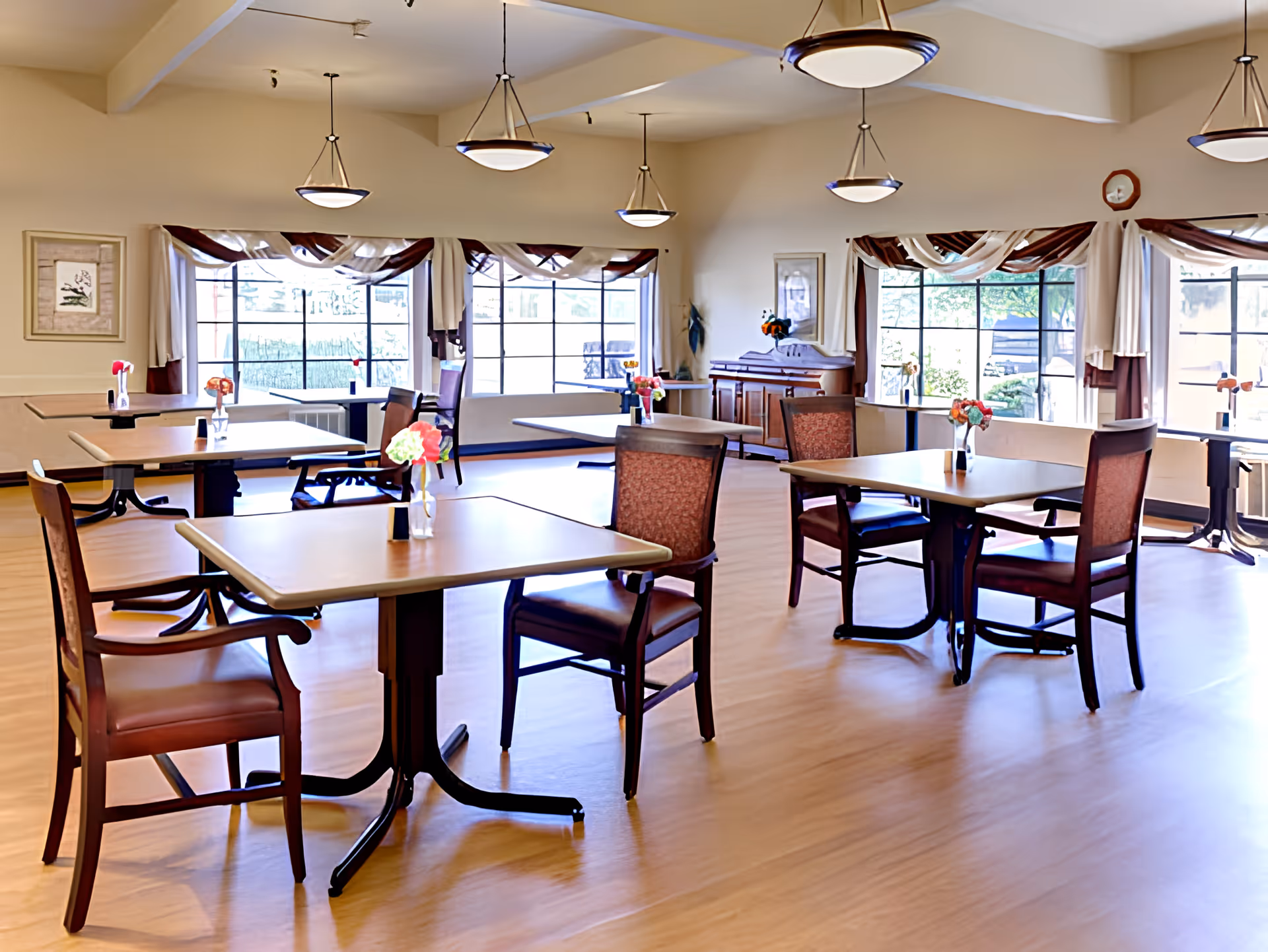 Bright dining room with multiple tables and chairs, vases of flowers, large windows and hanging pendant lights.