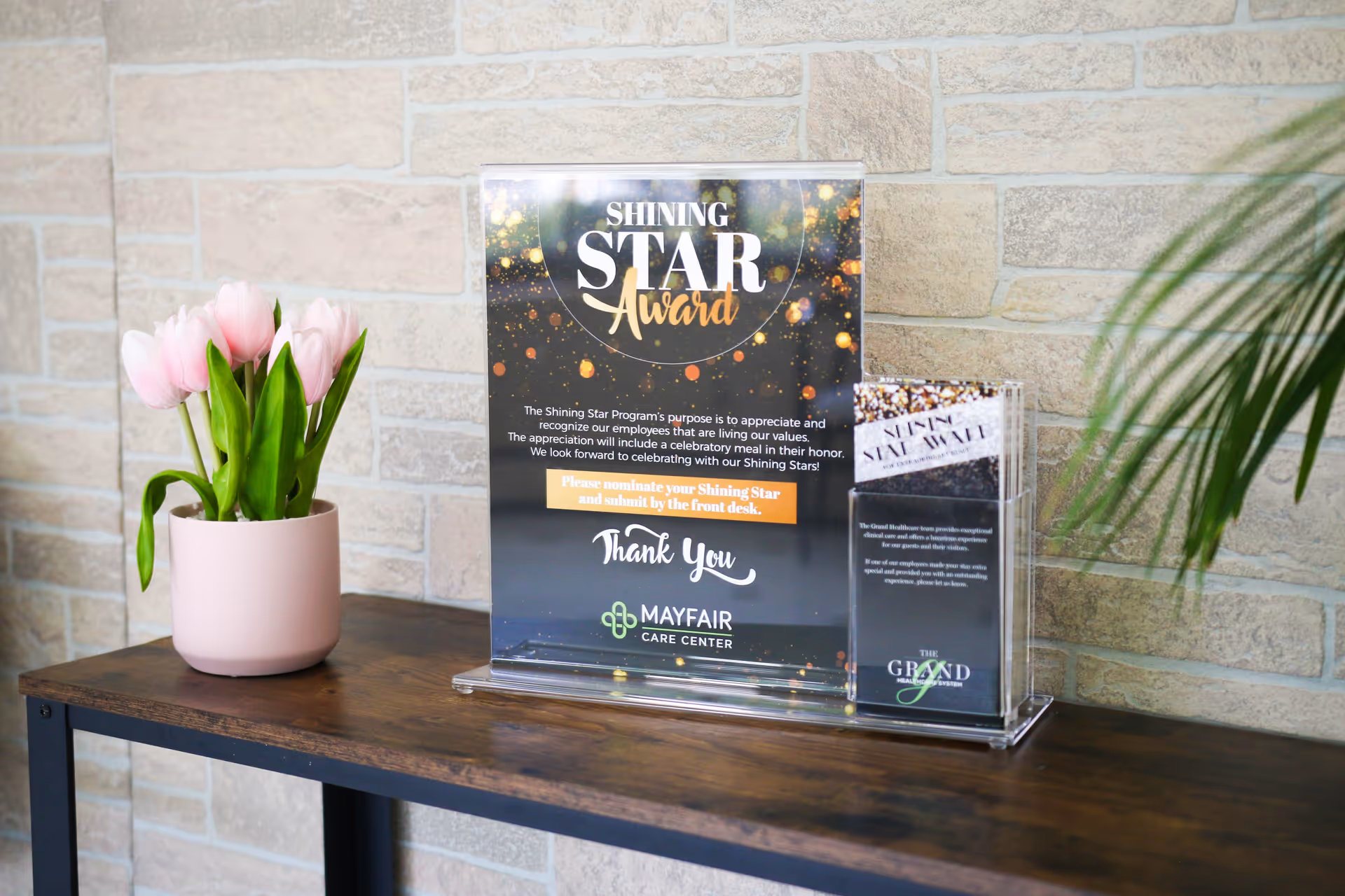 A wooden table against a light brick wall with a pink pot holding pink tulips on the left and a clear acrylic stand on the right displaying a Shining Star Award sign and brochures for Mayfair Care Center.
