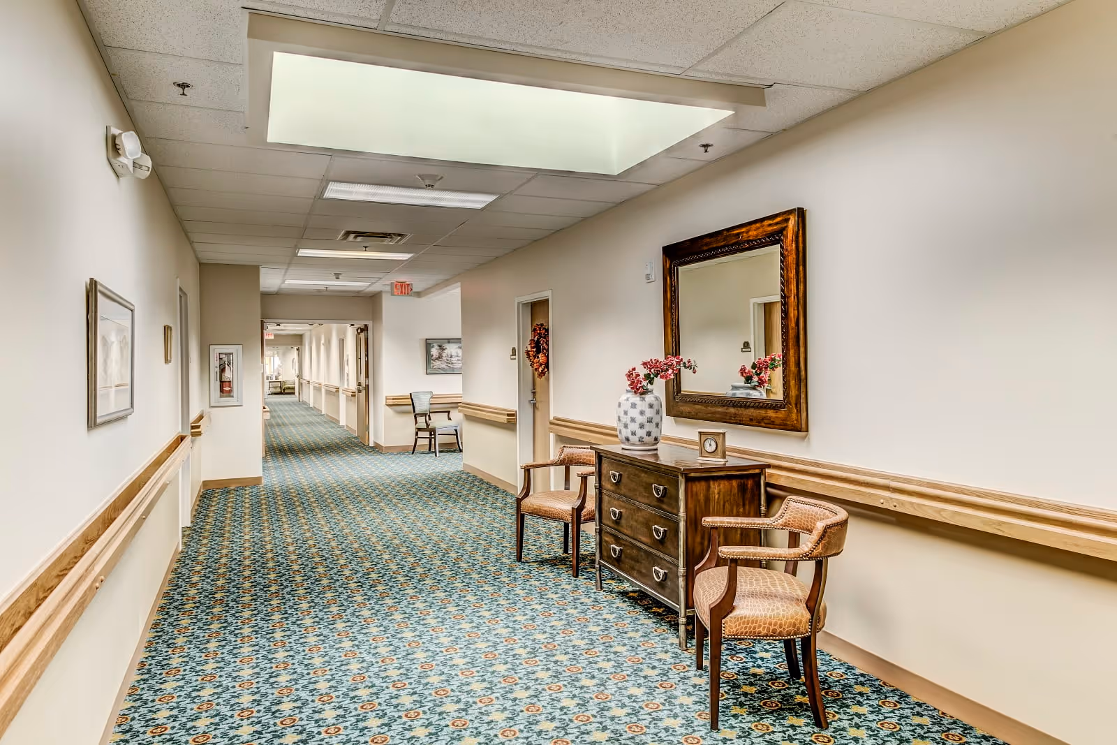 A long, well-lit hallway in a senior living facility with patterned carpet and beige walls. The hallway features wooden handrails on both sides, a wooden dresser with a vase of flowers and a clock on top, two wooden chairs with cushions, a large framed mirror above the dresser, and framed artwork on the walls. The ceiling has fluorescent lights and a skylight.