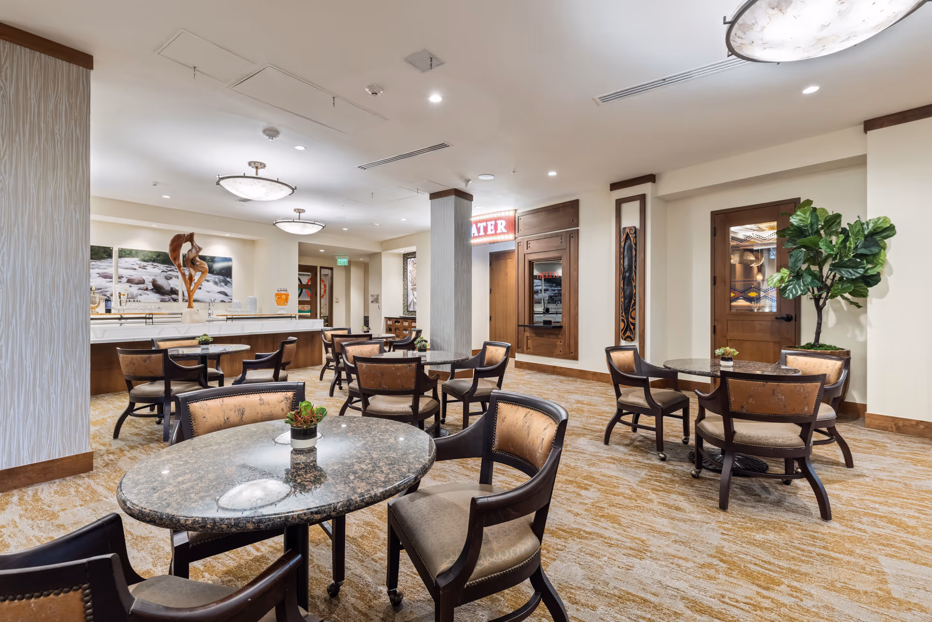 A spacious dining area with multiple round tables and cushioned chairs. The room features a carpeted floor, modern ceiling lights, and a large plant near a wooden door with stained glass. There is a counter with decorative items and artwork on the walls, creating a warm and inviting atmosphere.