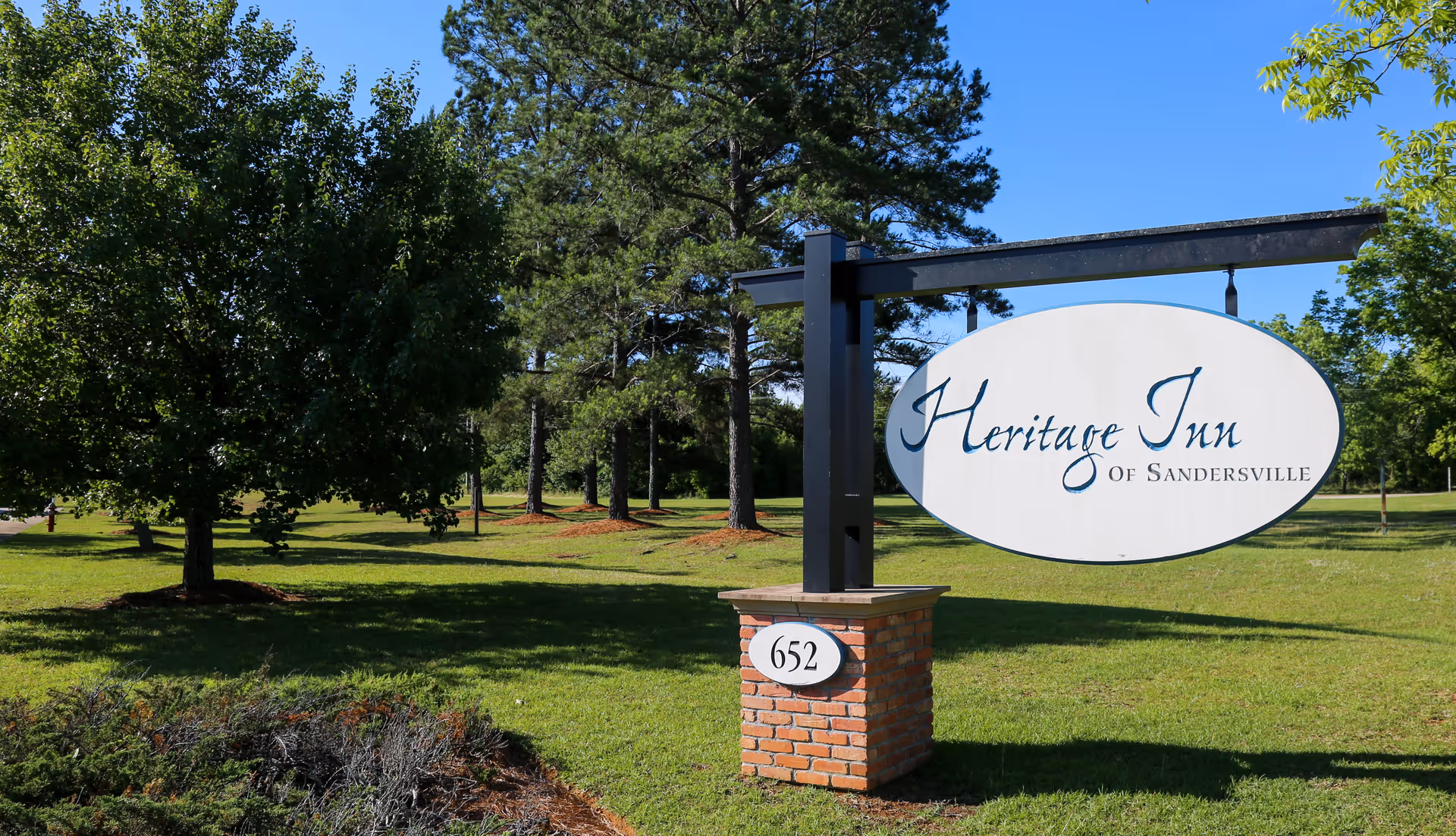 Outdoor view of a grassy area with trees and a sign that reads 'Heritage Inn of Sandersville' mounted on a black metal frame above a brick base with the number 652.