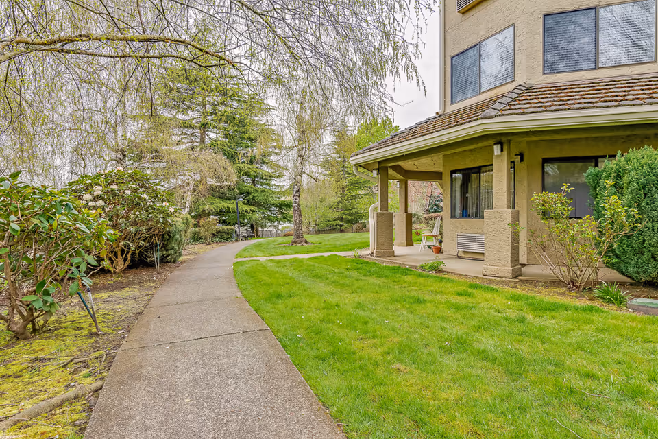 A paved walkway curves through a green lawn with bushes and trees on the left side. On the right side, there is a beige building with large windows and a covered patio area with columns. The scene appears to be outdoors with a cloudy sky.