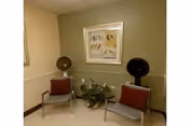 A small waiting or sitting area with two gray chairs, each with a red cushion. Between the chairs is a green potted plant. On the wall behind the chairs is a framed artwork featuring horses. The walls are painted in two tones, beige on the left and olive green on the right.