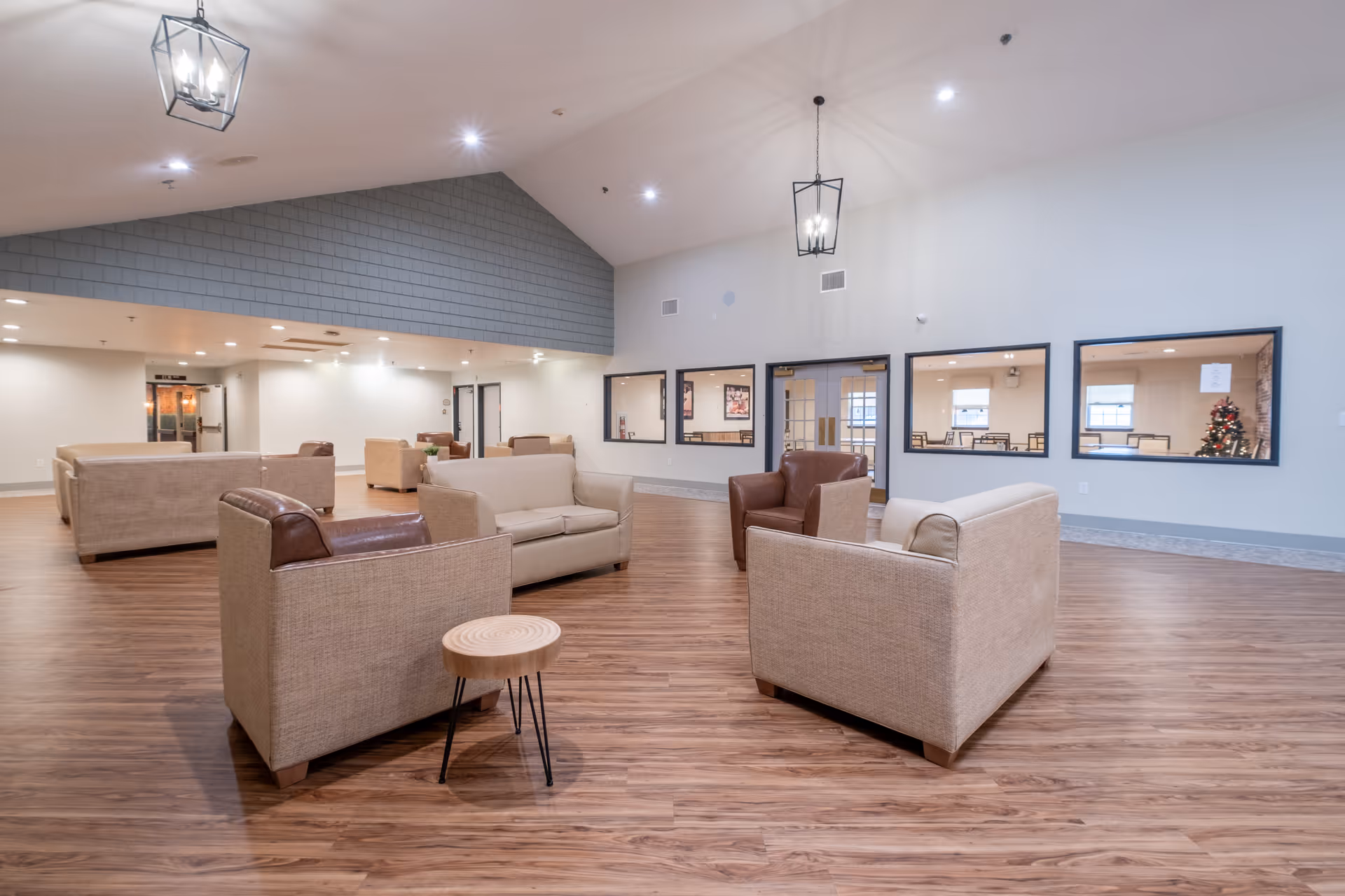 A spacious and well-lit common area with multiple beige and brown upholstered sofas and armchairs arranged on a wood floor. The room has a high vaulted ceiling with modern hanging light fixtures. Large windows and glass doors on one wall provide a view into an adjacent room with tables and chairs, and a decorated Christmas tree is visible in the background.