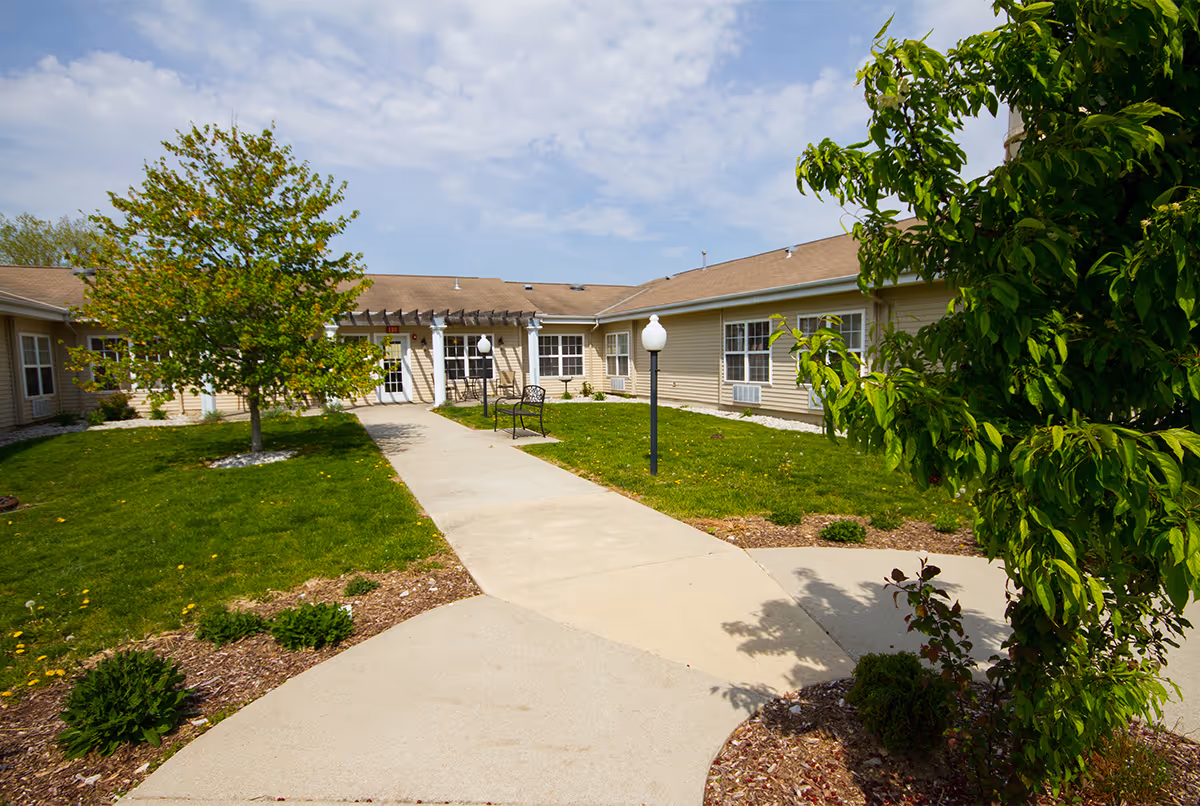 Outdoor courtyard area of a senior living facility with a concrete walkway, green grass, trees, benches, and a beige building with multiple windows under a partly cloudy sky.