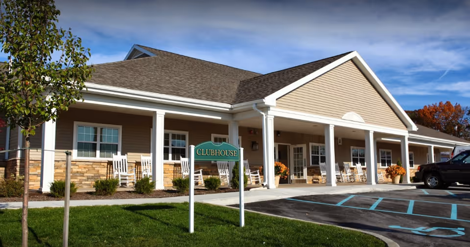 Front exterior of a single-story clubhouse with a covered porch, rocking chairs, a 'Clubhouse' sign and a parking area.