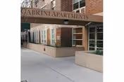 Exterior view of a brick building with a green awning that reads 'Cabrini Apartments' above the entrance. The sidewalk and part of the street are visible in front of the building.