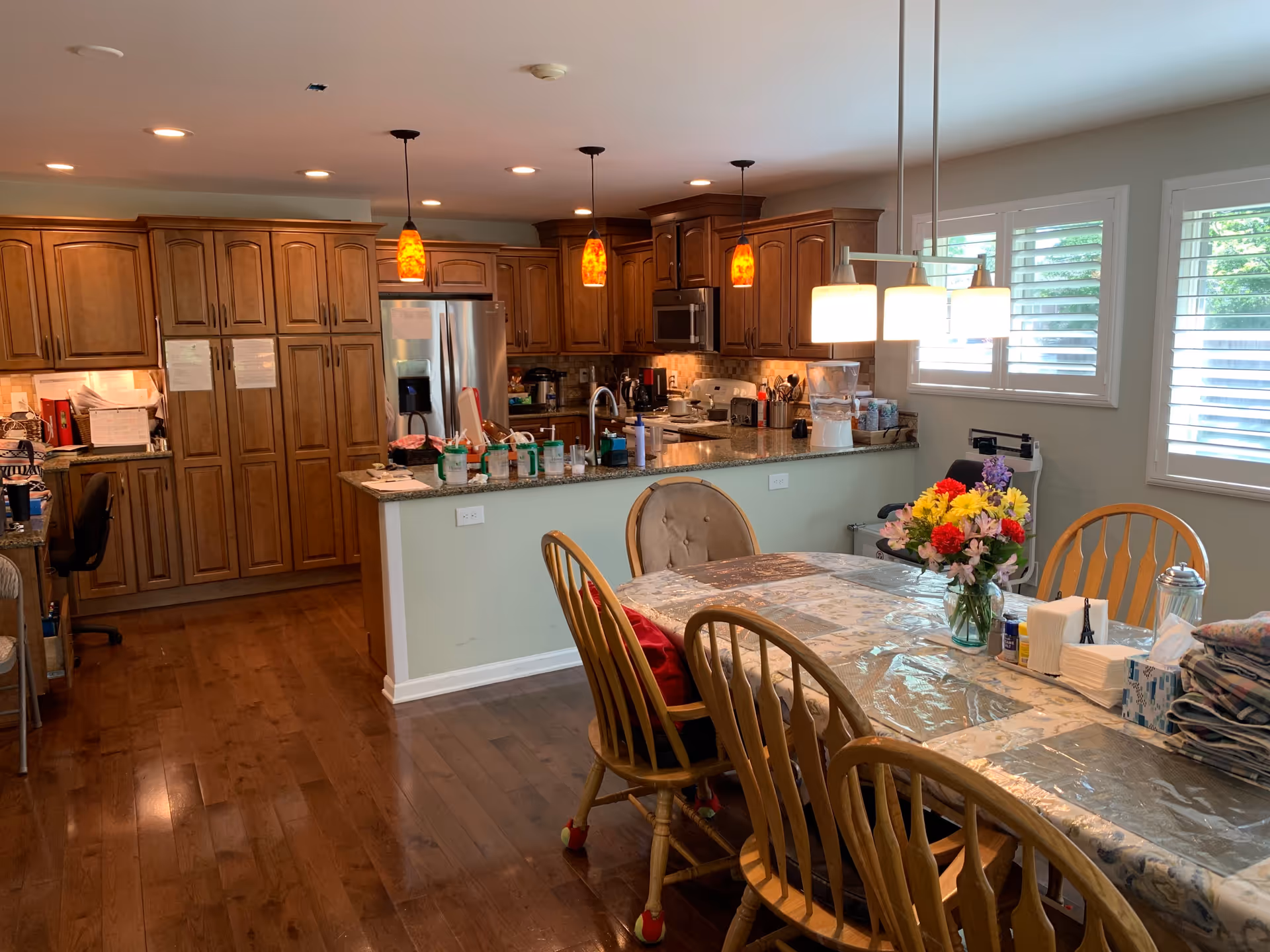 A spacious kitchen and dining area with wooden cabinets, stainless steel refrigerator, and granite countertops. The dining table is covered with a plastic tablecloth and has a vase of colorful flowers, napkins, and other items on it. Three pendant lights hang over the kitchen counter, and a modern light fixture hangs above the dining table. The room has wooden flooring and windows with white blinds letting in natural light.