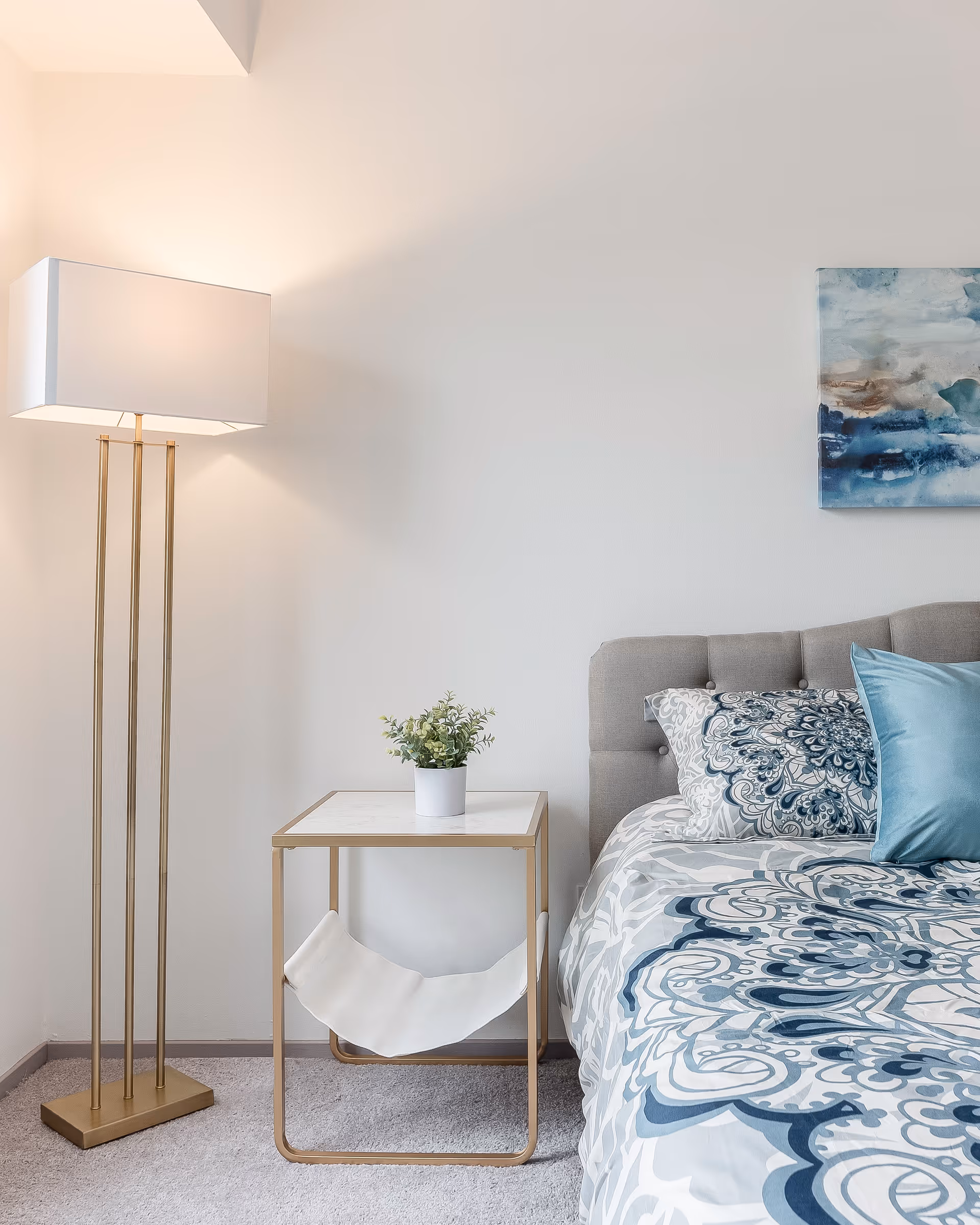 A cozy bedroom corner featuring a bed with a tufted gray headboard and blue patterned bedding, a small side table with a potted plant, and a tall floor lamp with a white rectangular shade.