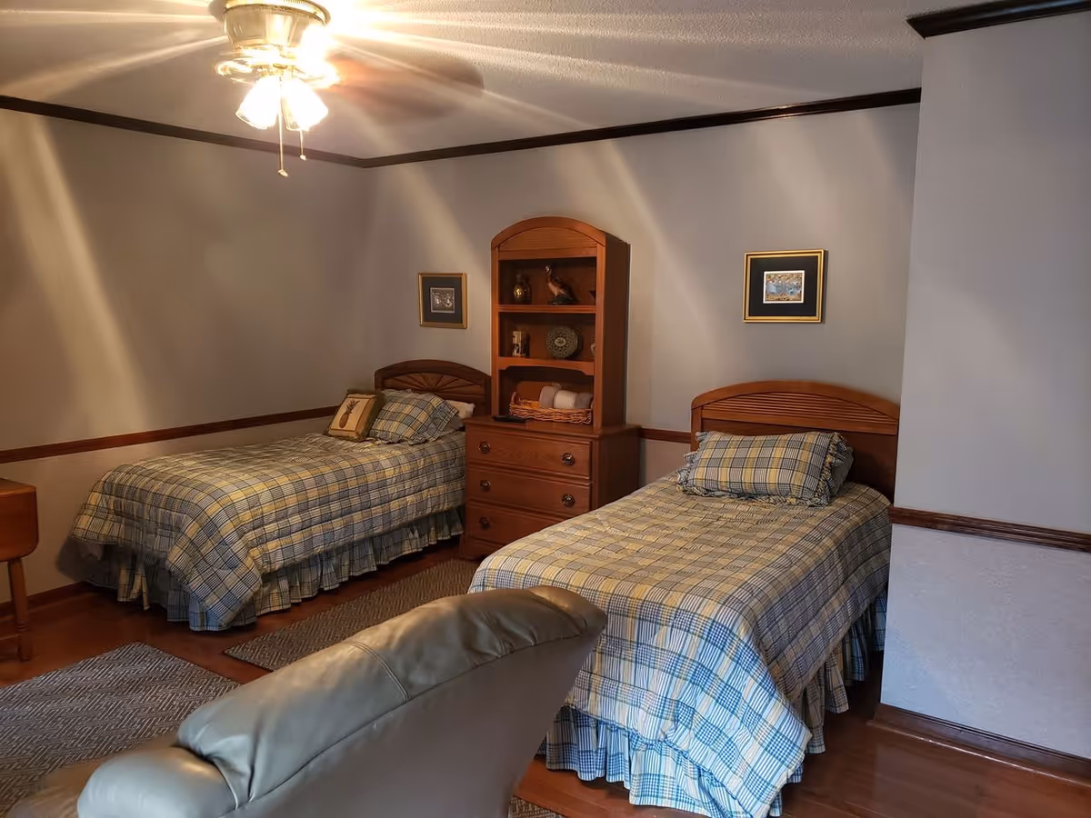 A bedroom with two single beds covered in matching plaid bedspreads. Between the beds is a wooden dresser with shelves holding decorative items. The room has wooden trim along the walls and a ceiling fan with lights. A beige recliner chair is partially visible in the foreground.