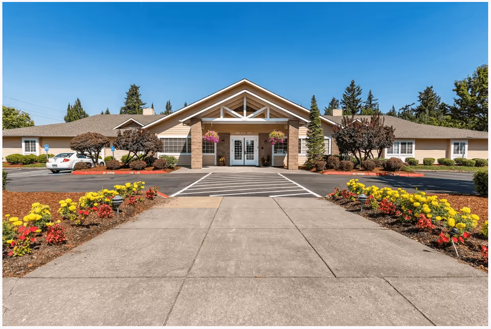 Front exterior view of a single-story senior living facility building with a peaked roof, beige siding, and a wide entrance with double doors. The entrance is flanked by hanging flower baskets and landscaped with trimmed bushes and colorful flowers along the walkway. There is a parking area on both sides of the entrance with a few cars parked and a clear blue sky above.